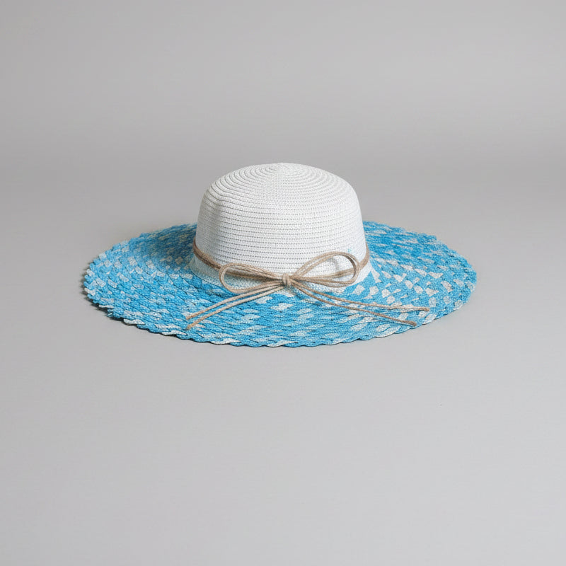 A white woven sun hat with a wide blue and white patterned brim and a beige twine bow, displayed on a brown cardboard box.