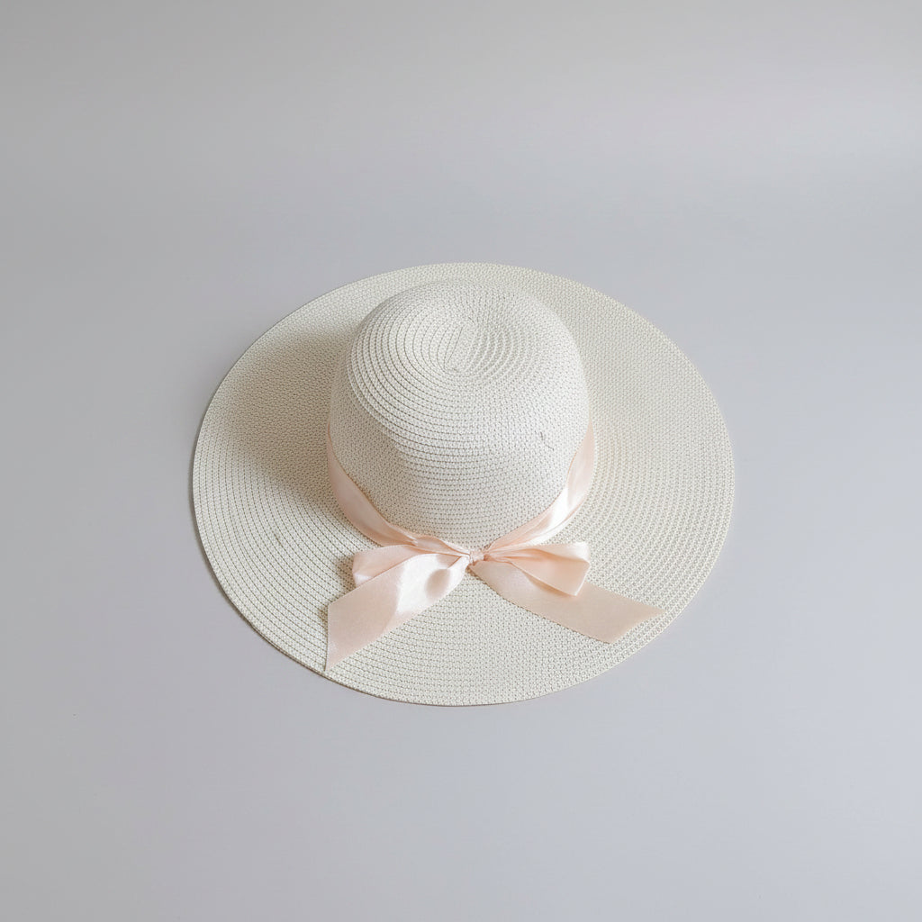 A white woven straw sun hat with a wide brim and a pale pink ribbon bow, resting on a brown cardboard box.