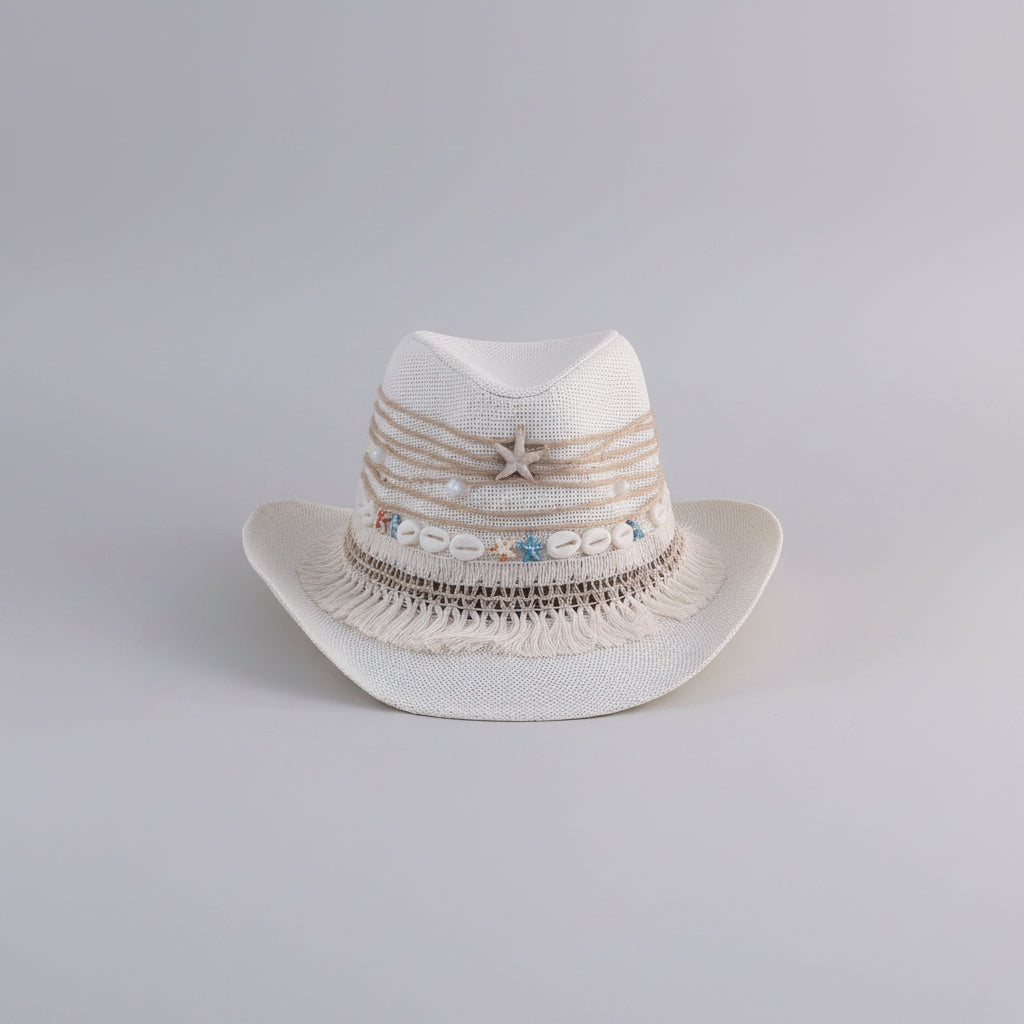 A white woven cowboy-style hat with beige rope trim, fringe, a starfish detail, and small blue and beige shell embellishments, displayed on a shelf in an indoor setting.