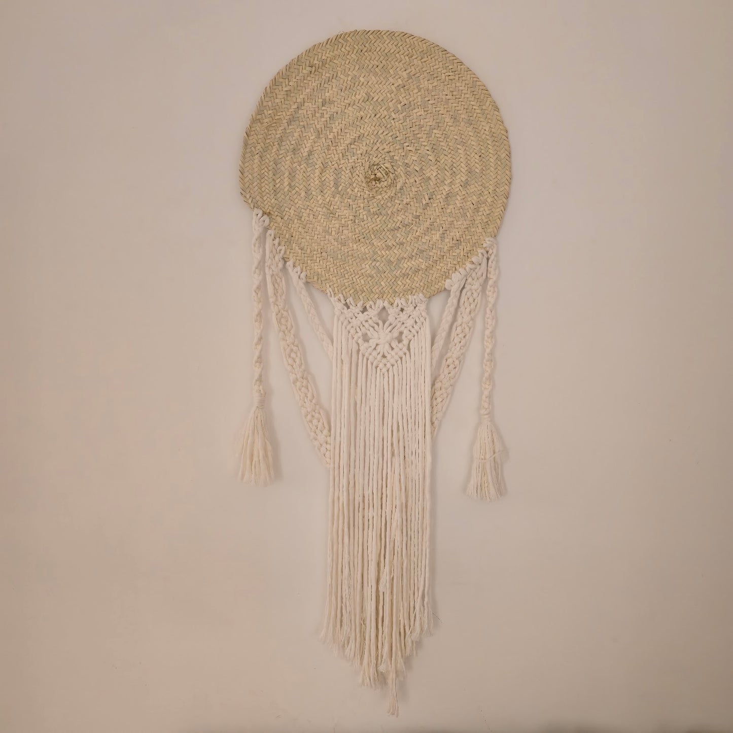 A circular woven wall hanging with a natural straw-colored round top and long cream macrame fringe tassels, displayed against a plain beige wall.