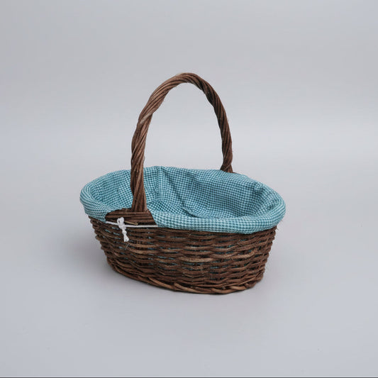 A brown woven basket with a twisted handle and a light blue and white gingham fabric lining, placed on a beige stone-tiled floor.