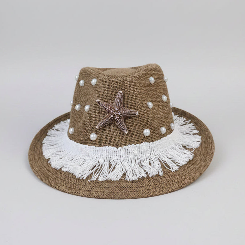 A brown woven fedora-style hat with white fringe, white pearl-like beads, a metallic starfish ornament, and a wide brim, photographed on a cardboard surface with a workshop or storage background.
