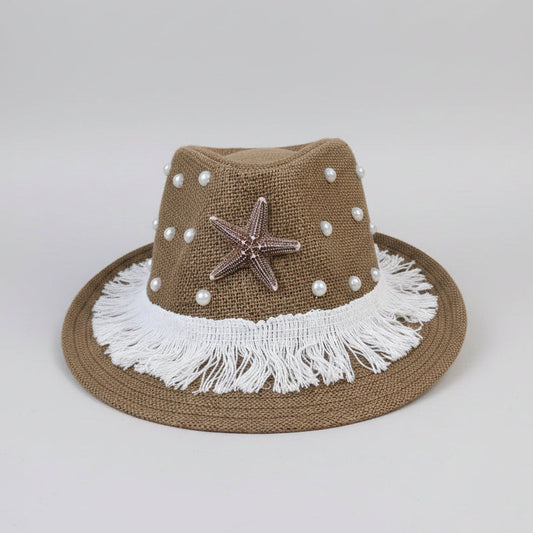 A brown woven fedora-style hat with white fringe, white pearl-like beads, a metallic starfish ornament, and a wide brim, photographed on a cardboard surface with a workshop or storage background.