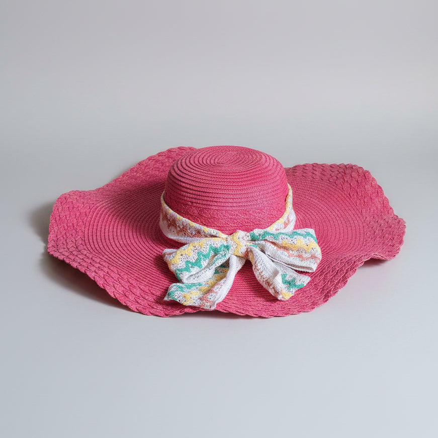 A bright pink woven sun hat with a wide brim and a multicolor floral bow, displayed on a brown cardboard box.
