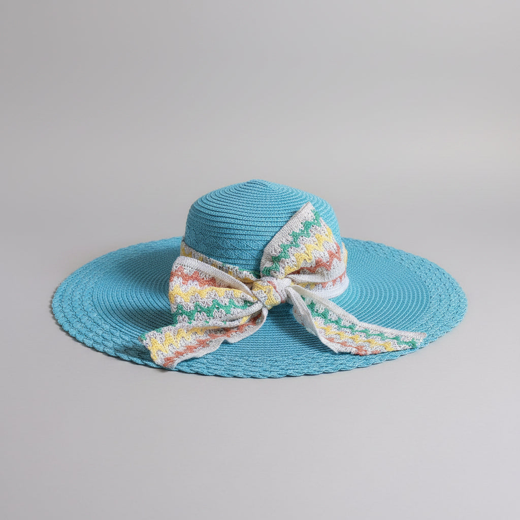 A bright turquoise blue woven sun hat with a wide brim and a large white bow featuring a multicolor zigzag pattern, displayed on a brown cardboard box.