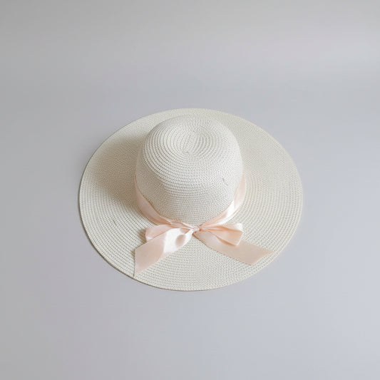A white woven straw sun hat with a wide brim and a pale pink ribbon bow, resting on a brown cardboard box.