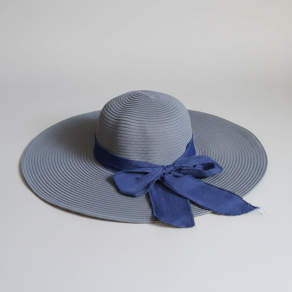 A wide-brimmed straw sun hat in a muted blue-gray color, featuring a dark blue satin ribbon tied in a bow at the crown, displayed on a brown cardboard box.