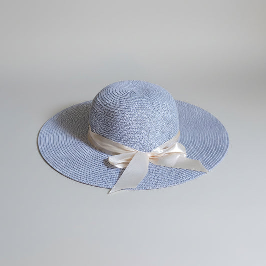 A light blue woven sun hat with a cream satin ribbon bow, resting on a brown cardboard box.