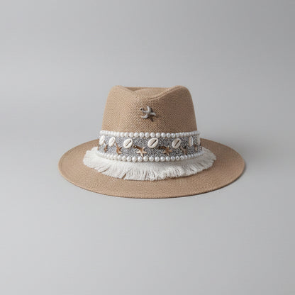 A light brown woven fedora hat with a white beaded band and fringe trim, displayed on a white shelf in a home setting.