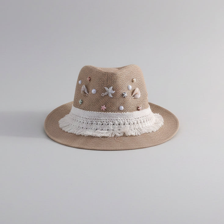 A beige woven fedora hat with white lace trim and decorative pastel beads and charms, displayed on a white shelf in a home setting.