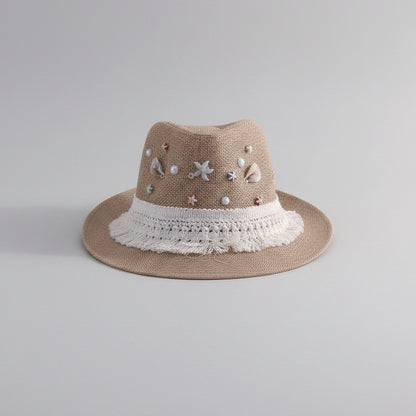 A beige woven fedora hat with white lace trim and decorative pastel beads and charms, displayed on a white shelf in a home setting.