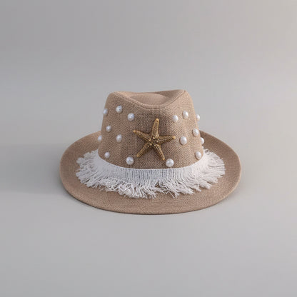 A beige straw fedora hat with white faux pearls, a gold starfish ornament, and a white frayed trim, displayed on a white shelf.