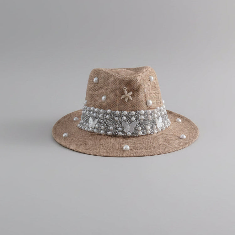 A beige straw fedora hat with a wide brim, decorated with a white pearl band featuring butterfly embellishments, a small silver flower charm, and scattered white pearls, displayed on a light gray shelf.