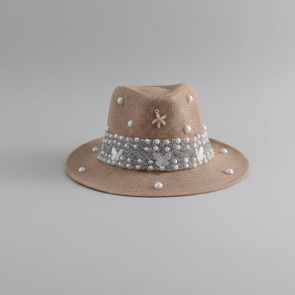 A beige straw fedora hat with a wide brim, decorated with a white pearl band featuring butterfly embellishments, a small silver flower charm, and scattered white pearls, displayed on a light gray shelf.