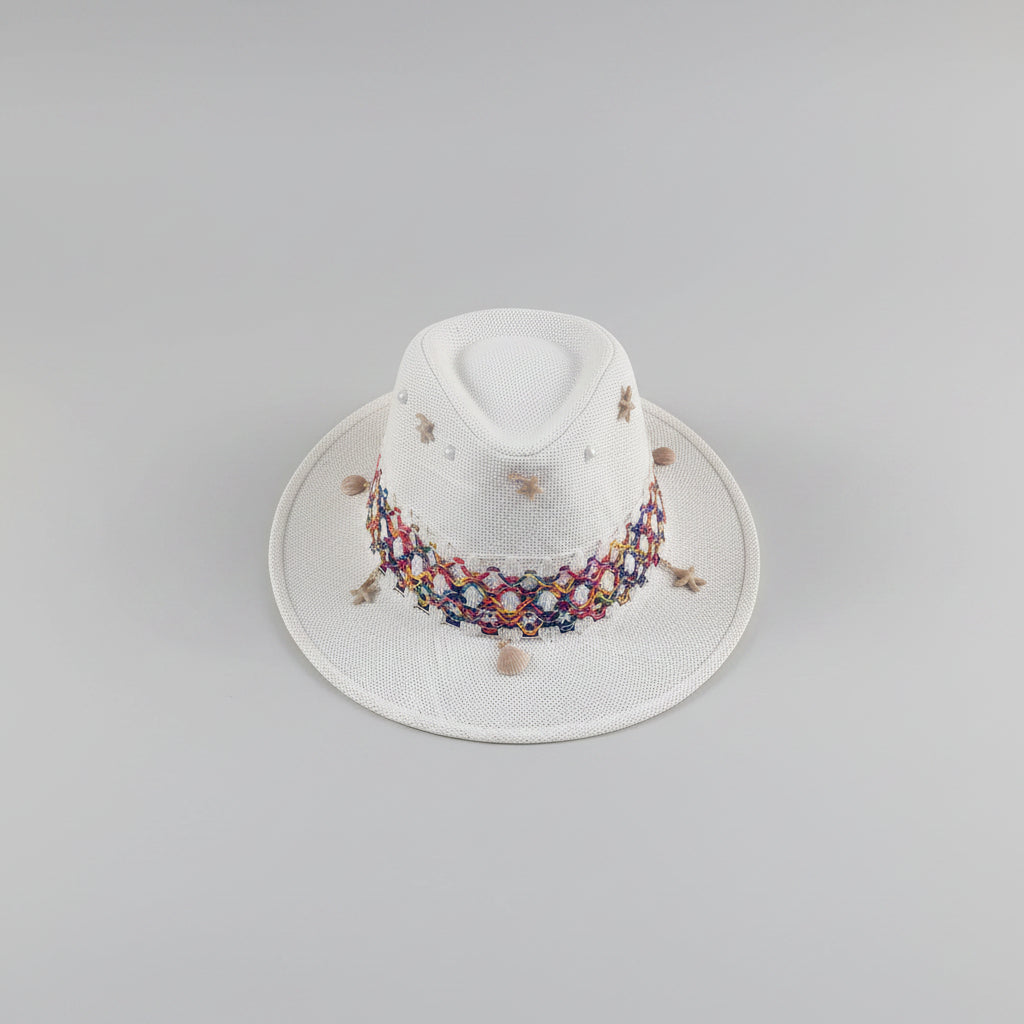 A white woven fedora hat with a colorful embroidered band and small decorative beads, resting on a light gray shelf in an indoor setting.