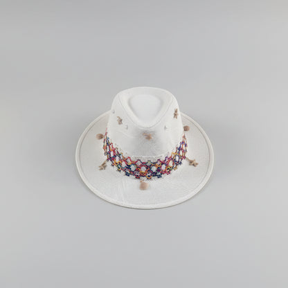 A white woven fedora hat with a colorful embroidered band and small decorative beads, resting on a light gray shelf in an indoor setting.