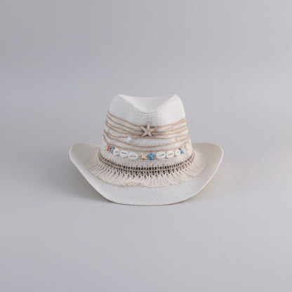 A white woven cowboy-style hat with beige rope trim, fringe, a starfish detail, and small blue and beige shell embellishments, displayed on a shelf in an indoor setting.
