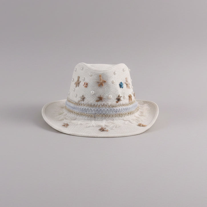 A white woven fedora-style hat with embroidered star details and a decorative band, resting on a light gray shelf in a styled indoor setting.