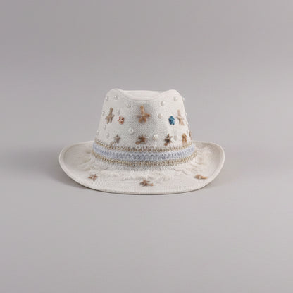 A white woven fedora-style hat with embroidered star details and a decorative band, resting on a light gray shelf in a styled indoor setting.