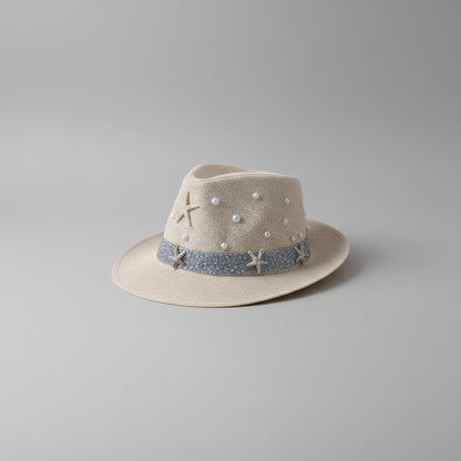 A light beige woven fedora hat with a blue glittery band, decorated with white pearl-like beads, a starfish, and a small anchor, displayed on a shelf.