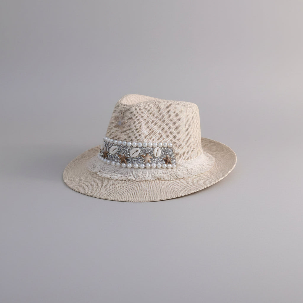 A light beige straw fedora hat with a decorative band featuring faux pearls, metallic shells, and a small star charm, displayed on a light-colored shelf.
