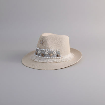 A light beige straw fedora hat with a decorative band featuring faux pearls, metallic shells, and a small star charm, displayed on a light-colored shelf.