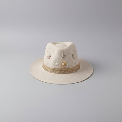 A light beige woven fedora hat with decorative embroidered flowers, pearl-like beads, and a patterned band, resting on a shelf in a styled indoor setting.