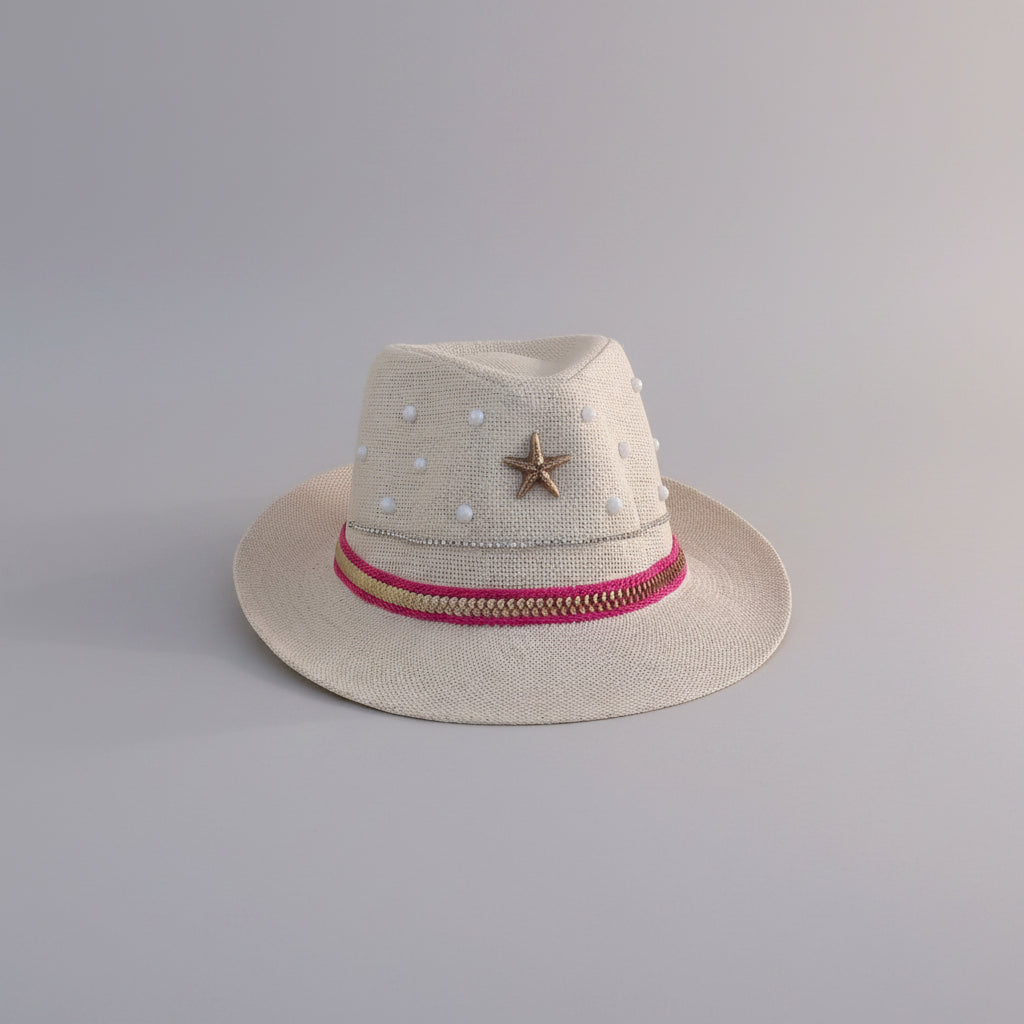 A light beige straw fedora hat with white pearl-like embellishments, a brown starfish accent, and a pink and gold band, displayed on a light gray shelf.