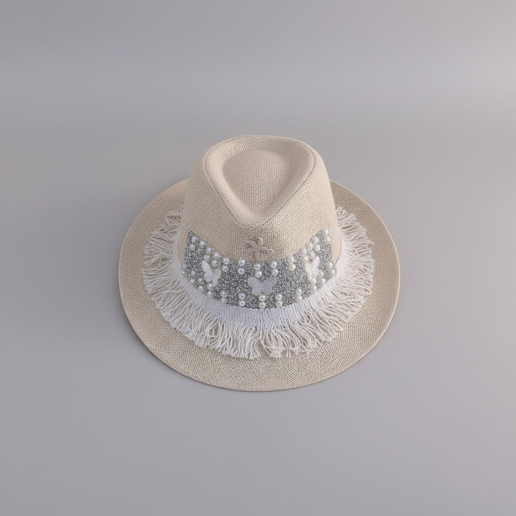 A light beige straw fedora hat with a decorative band of white pearls, silver rhinestones, and white fringe trim, displayed on a white shelf.