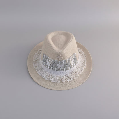 A light beige straw fedora hat with a decorative band of white pearls, silver rhinestones, and white fringe trim, displayed on a white shelf.