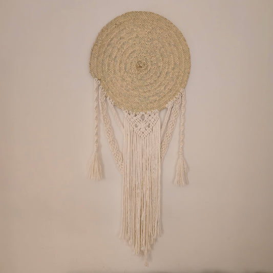 A circular woven wall hanging with a natural straw-colored round top and long cream macrame fringe tassels, displayed against a plain beige wall.