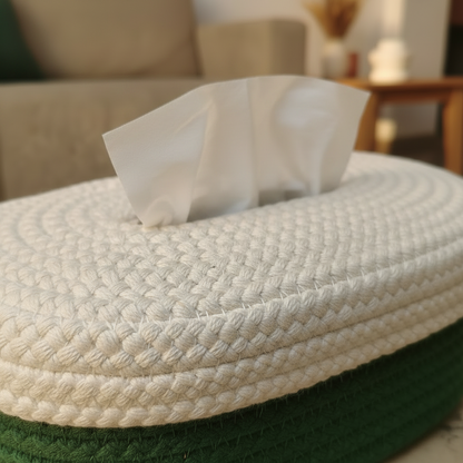 A close-up of a woven rope tissue box cover in off-white with a dark green base, with a white tissue paper protruding from the top, set against a softly blurred living room background.