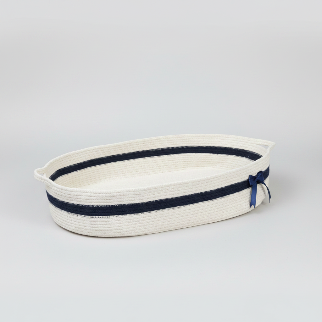 An oval-shaped woven rope basket in off-white with a navy blue stripe and a matching navy ribbon bow on the side, photographed on a seamless light gray studio background.