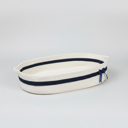 An oval-shaped woven rope basket in off-white with a navy blue stripe and a matching navy ribbon bow on the side, photographed on a seamless light gray studio background.