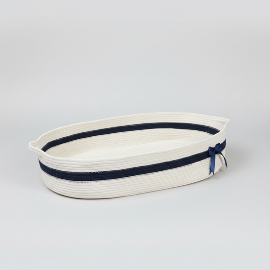 An oval-shaped woven rope basket in off-white with a navy blue stripe and a matching navy ribbon bow on the side, photographed on a seamless light gray studio background.