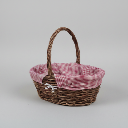 A small oval wicker basket with a brown woven body, a single curved handle, and a pink-and-white gingham fabric liner tied at the front, photographed on a light gray studio background.