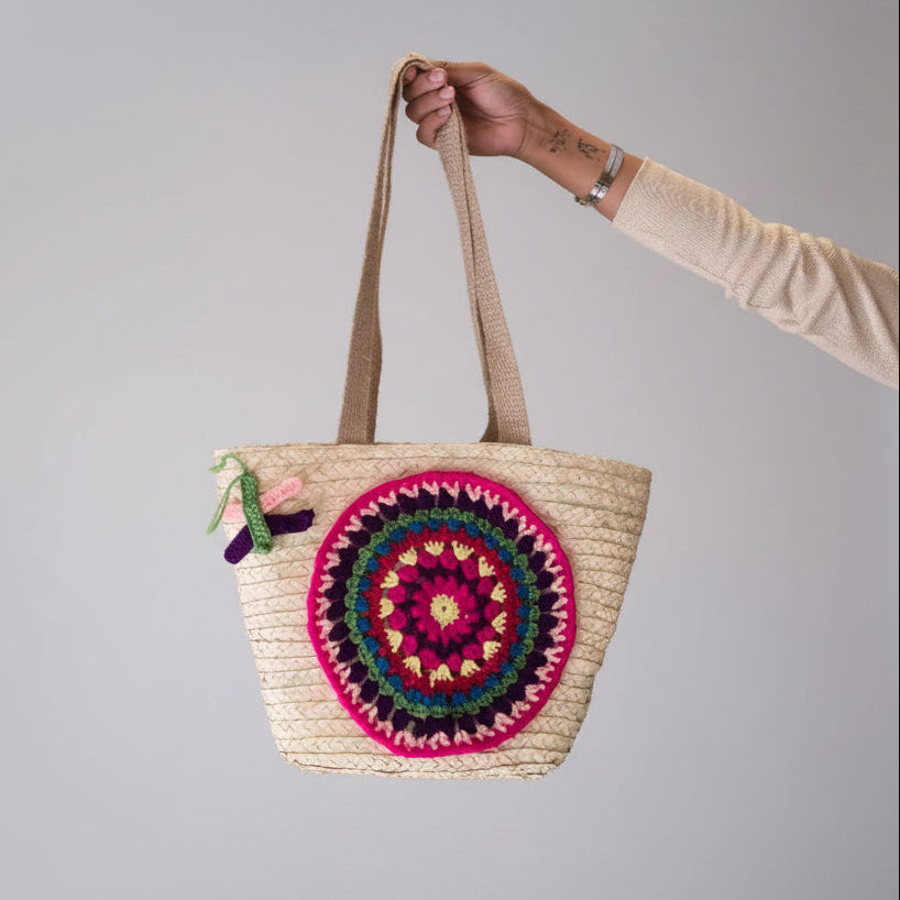 A straw tote bag with a colorful crocheted circular design on the front, held by a person's arm against a plain gray background.