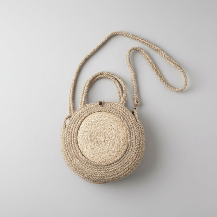 A round woven straw handbag with a braided rope handle and shoulder strap, resting on a white fabric surface.