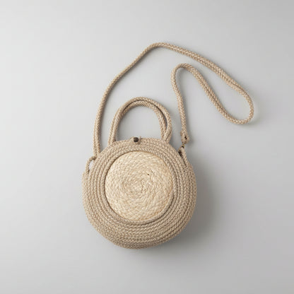 A round woven straw handbag with a braided rope handle and shoulder strap, resting on a white fabric surface.