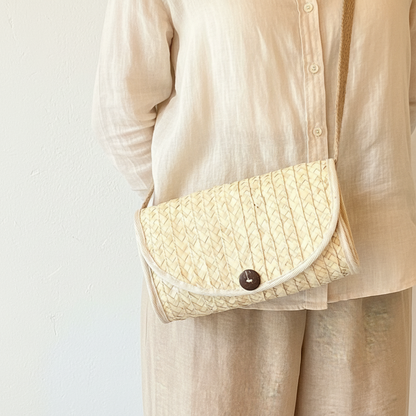 A woven straw clutch bag with a dark brown button closure and a beige shoulder strap, worn crossbody by a person wearing a light beige linen button-up shirt and matching trousers.