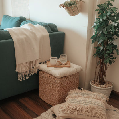 A woven natural-fiber ottoman or pouf with a cream cushion, styled in a cozy room corner beside a teal sofa with a cream throw blanket. A small wooden tray holding a white mug and a clear glass sits on top, with a hanging basket and potted plant nearby.