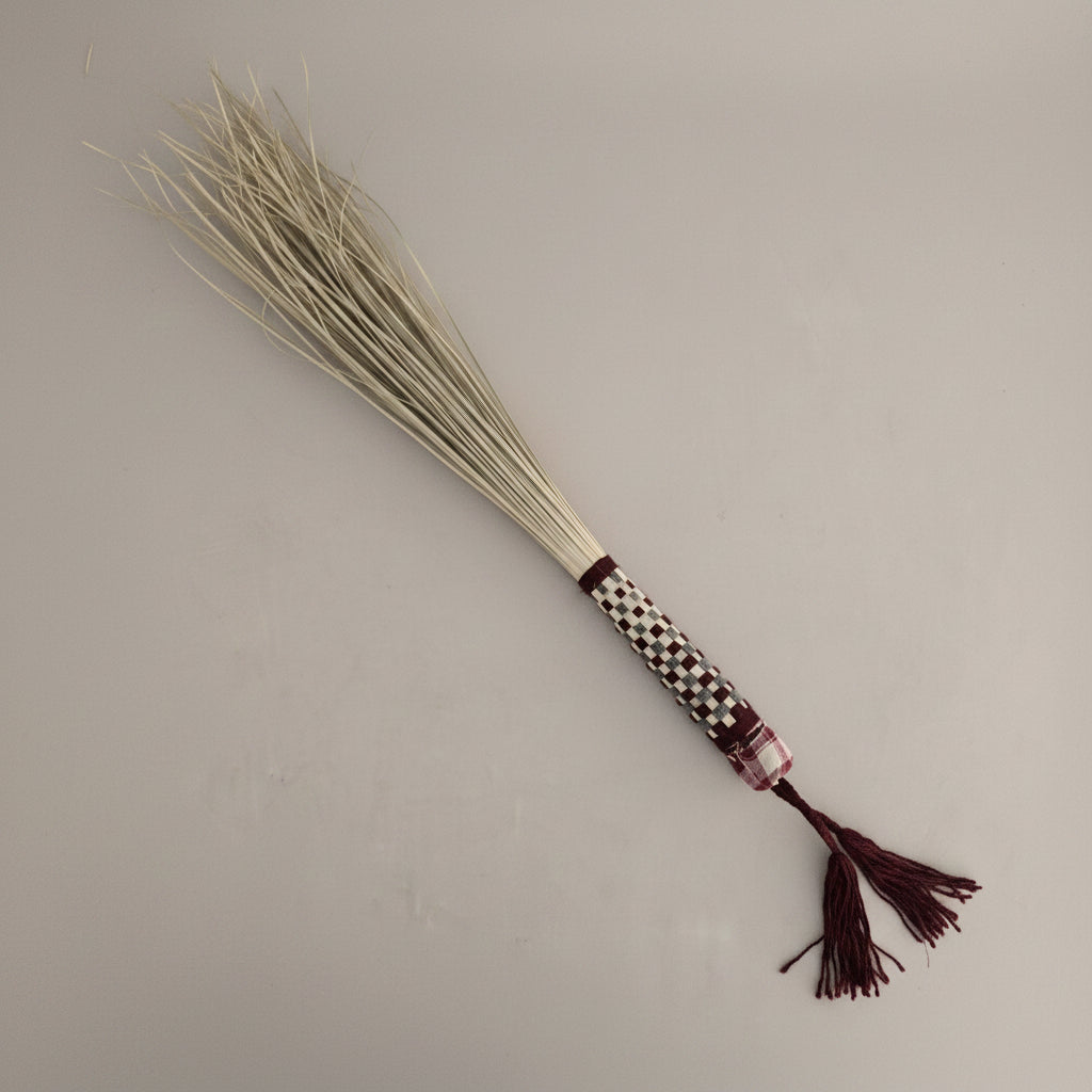 Karkadeh manasha palm hand broom with burgundy woven handle and dark red tassel, on a plain studio backdrop — Khoos