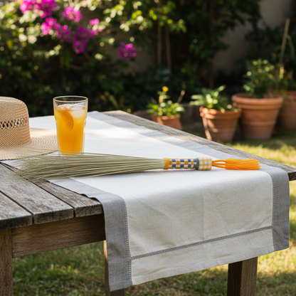 Yostafandi manasha palm broom with gold-and-black gingham handle and bright orange tassel on a cream linen tablecloth in a sunlit garden, beside a tall glass of chilled bright orange Yostafandi mandarin drink with ice — Khoos