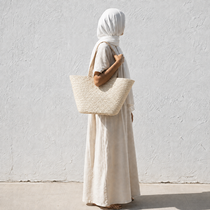 A woman in a white headscarf, beige tunic, and long skirt stands in profile against a textured white wall, carrying a large woven straw tote bag.