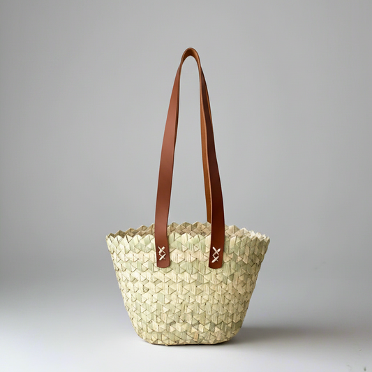 Wavy handwoven palm tote with two tan leather handles, shown alone on a grey studio backdrop — Khoos